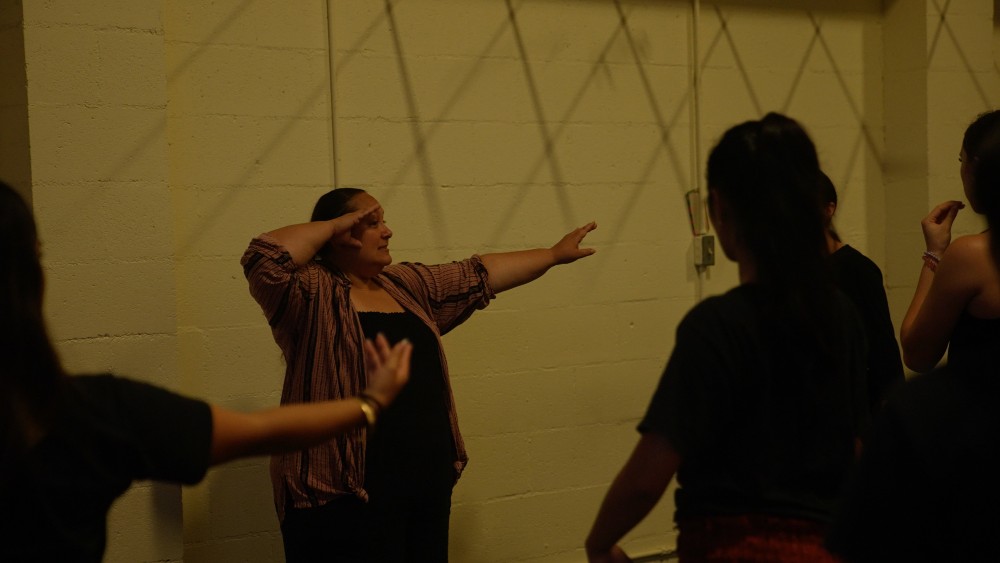 A person stands in front of a room of people with one arm raised up in front of her head and another outstretched to the side during a hula