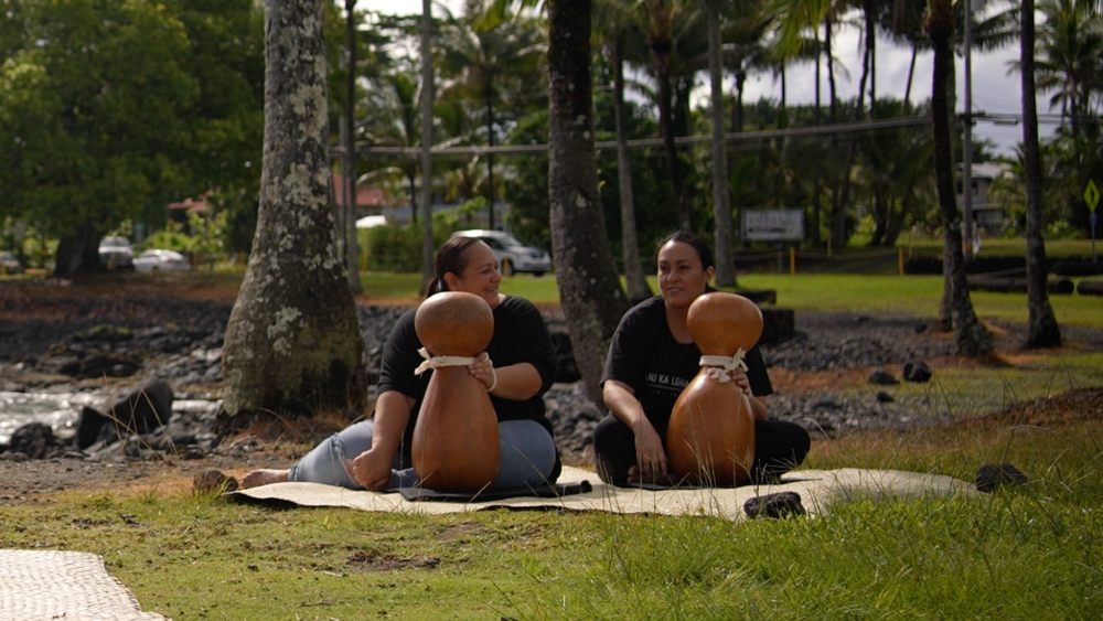 Two female hula practitioners performing a mele on the grass at the beach.