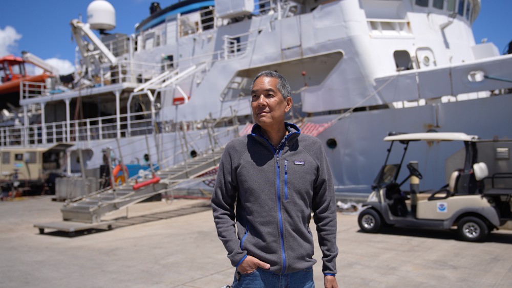 A person stands in front of a research vessel prior to a science expedition