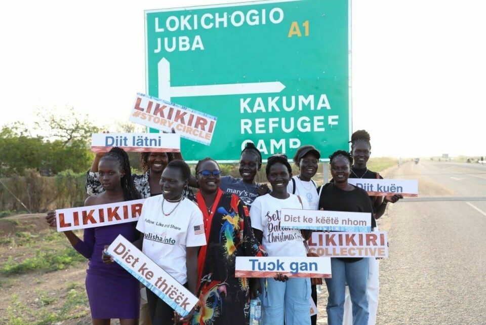 A group of women are standing in front of a street sign showing the way to Juba, South Sudan and Kakuma Refugee Camp, Kenya.